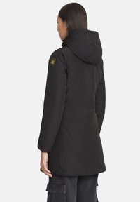 Parka nera isolata con cappuccio, dal taglio aderente, tessuto strutturato e toppa del logo gialla sul braccio sinistro.