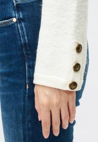 Punho de um suéter branco e texturizado apresenta três botões decorativos em tom dourado, combinado com um denim azul escuro, enfatizando o contraste nos materiais e cores.