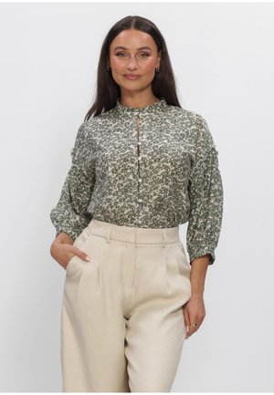 Femme aux longs cheveux foncés portant une blouse verte à fleurs boutonnée et un pantalon beige taille haute, debout avec une main dans la poche.