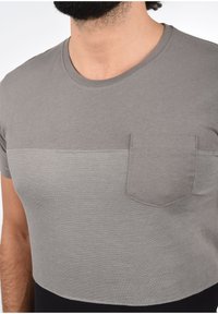 Camiseta gris de manga corta en una tela suave y texturada, con una costura horizontal y un pequeño bolsillo en el pecho. Escote redondo y diseño corto.