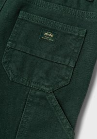Tasca in denim verde scuro con una finitura strutturata. Presenta un'etichetta rettangolare con testo dorato che recita "Denim Originale dal 1986".