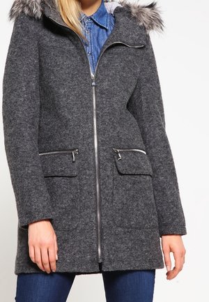 Femme portant un manteau en laine gris foncé avec capuche bordée de fourrure, fermeture éclair argentée à l'avant, poches zippées et à rabat, par-dessus une chemise en denim et un jean bleu.