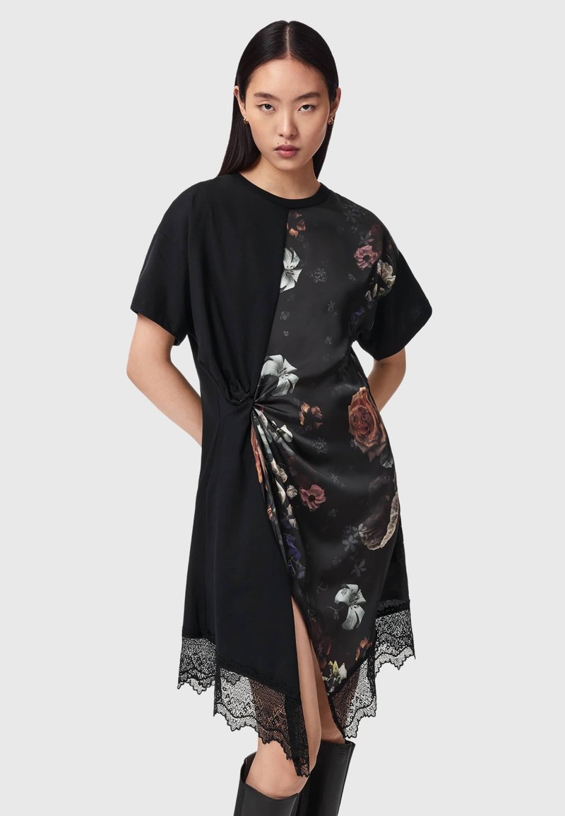 Femme portant une robe noire mi-longue avec un panneau en satin floral, un détail froncé à la taille, un bord en dentelle et des manches courtes, debout devant un fond gris.