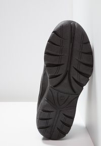 Semelle d'une chaussure noire avec des rainures profondes et un motif de bande de roulement texturé, reposant verticalement contre un mur blanc et un sol blanc.