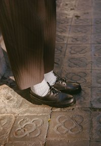Chaussures en cuir noir avec des lacets, associées à des chaussettes blanches à côtes, portées avec un pantalon marron à rayures, sur un fond en pierre texturée.