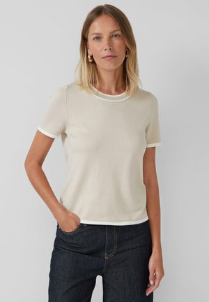 Femme portant un haut en tricot à manches courtes beige avec des bordures blanches, un jean bleu foncé et des boucles d'oreilles dorées en forme de cerceau, se tenant contre un fond clair uniforme.
