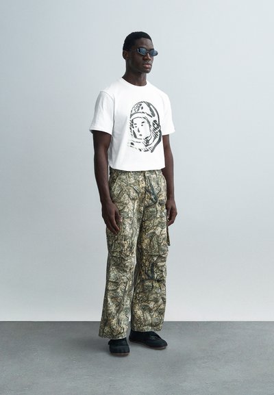 Λευκό κοντό t-shirt με γραφικό σχέδιο, συνδυασμένο με παραλλαγμένα cargo παντελόνια σε γήινους τόνους. Μαύρα γυαλιά ηλίου και υποδήματα ολοκληρώνουν την εμφάνιση.