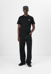 Zwarte katoenen t-shirt met een klein logo, gecombineerd met losse zwarte spijkerbroek en zwarte schoenen; drager heeft een grijze pet.