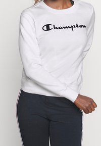 Kvinna klädd i en vit Champion-tröja och marinblå byxor med röda och vita sidostripes, stående med höger arm böjd.