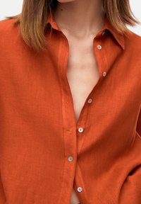 Chemise en lin couleur rouille avec un front à boutons, dotée d'un col classique, d'une texture subtile et de boutons de couleur claire.