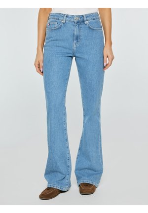 Osoba nosząca jasnoniebieskie, rozkloszowane dżinsy z wysokim stanem i brązowe zamszowe buty, stojąca na tle jasnego, gładkiego tła.