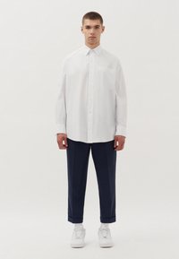 Witte lange mouw shirt met verticale strepen, knoopsluiting aan de voorkant, enkele borstzak, gecombineerd met marineblauwe broek en witte sneakers.