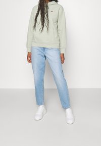 Jasnozielona bluza z kapturem i kieszenią kangurka, w połączeniu z jasnoniebieskimi dżinsami o prostych nogawkach oraz białymi sneakersami na platformie.