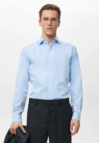 Svetlo modra srajca z dolgimi rokavi, z ovratnikom in gumbno zapiralo, kombinirana s temnimi hlačami. Gladek material in prilagojen kroj poudarjata obliko.