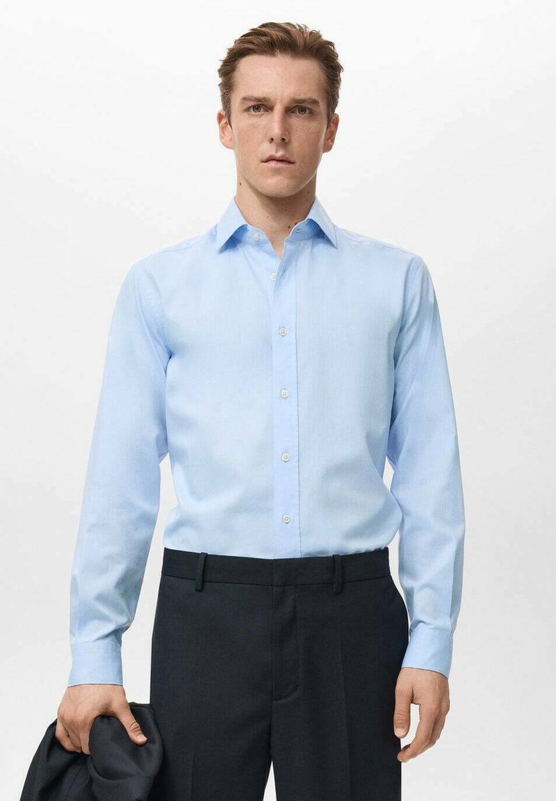 Svetlo modra srajca z dolgimi rokavi, z ovratnikom in gumbno zapiralo, kombinirana s temnimi hlačami. Gladek material in prilagojen kroj poudarjata obliko.