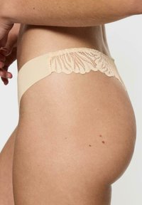 Beżowe koronkowe stringi z gładkim, elastycznym pasem i falbaniastym wykończeniem koronkowym z tyłu, charakteryzujące się minimalistycznym wzornictwem.