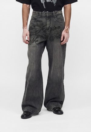 Személy széles szárú, kifakult fekete farmert visel, melyen fekete dőlt betűs szöveg található a comb felső részén, valamint fényes fekete cipőket egyszerű háttér előtt.