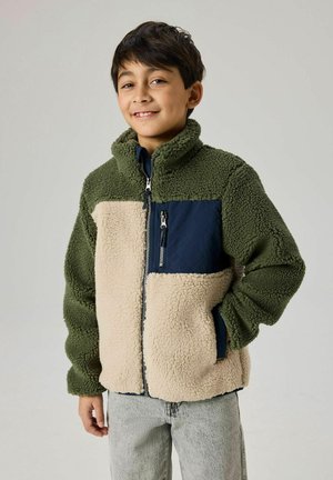 Ragazzo che indossa una giacca in pile verde e beige con una tasca sul petto con cerniera blu navy, sorridente con una mano in tasca.
