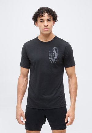 Νέος άνδρας με σγουρά μαλλιά στέκεται με το πρόσωπο προς τα εμπρός, φορώντας μαύρο μπλουζάκι με αφηρημένο σχέδιο και μαύρα σορτς σε λευκό φόντο.