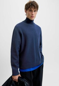 Jersey de cuello alto azul oscuro hecho de material suave con cuello acanalado, llevado sobre una camisa azul, combinado con pantalones de rayas.