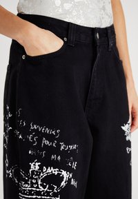 Un pantalon en denim noir avec une coupe décontractée qui présente des textes manuscrits blancs et des designs graphiques sur les jambes. Comprend une poche latérale et du matériel en métal.