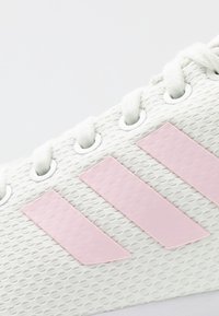 Vita mesh-sneakers med tre rosa gummistreck, runda vita öljetter och strukturerade skosnören. Närbild som framhäver detaljer och material.