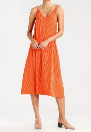 Femme portant une robe midi orange sans manches avec de fines rayures verticales et des chaussures beige à talons bas, debout les mains jointes.