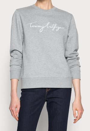 Persoon draagt een lichtgrijze trui met "Tommy Hilfiger" in witte cursief over de borst, gecombineerd met donkerblauwe spijkerbroek.