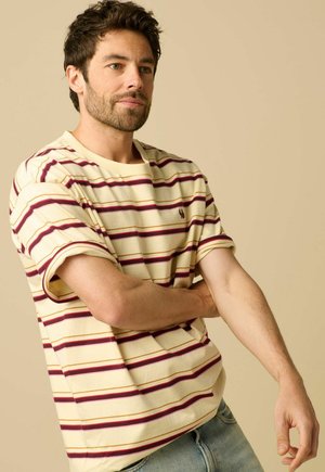 Homme aux cheveux foncés et à la barbe, portant un t-shirt rayé jaune, bordeaux et rouge, remontant sa manche gauche devant un fond beige.