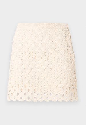 Γυναίκα όρθια με τα χέρια πίσω από την πλάτη, φορώντας ένα αμάνικο, ανοιχτό κίτρινο τοπ, μια κρεμ δαντελωτή μίνι φούστα και μαύρα παπούτσια Mary Jane.