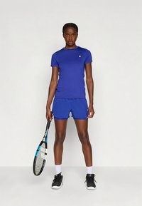 Γυναικείο αθλητικό ντύσιμο σε βασιλικό μπλε, που περιλαμβάνει ένα εφαρμοστό T-shirt και σορτς που ταιριάζουν. Περιλαμβάνει ρακέτα τένις και μαύρα αθλητικά παπούτσια.