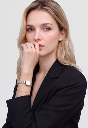 Szőke nő fekete blézerben, gyöngyfényű fülbevalóval, ezüst és arany órát, finom karkötőt, valamint aranygyűrűt visel, keze az ajkai közelében.
