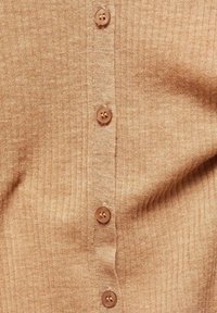 Světle hnědý žebrovaný cardigan s pěti kulatými knoflíky podél zapínání. Měkká textura s jemnou elastickou a splývavou strukturou.