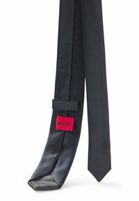 Cravate en satin noir avec une extrémité pointue et une surface texturée. Comprend une étiquette rouge "HUGO" et un accent rouge près de la boucle.