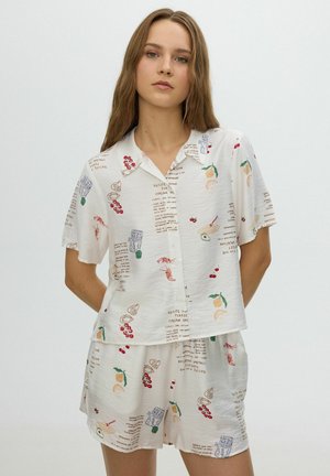 Joven mujer vestida con camisa blanca de manga corta y pantalones cortos con estampado de frutas coloridas y texto, de pie con las manos detrás de la espalda contra un fondo liso.
