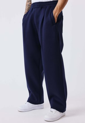 Personne portant un pantalon de survêtement bleu marine ample et des baskets blanches, debout les mains dans les poches devant un fond uni.