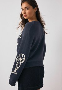 Mørkeblå fleece-trøje med hvid blomsterbroderi på ærmerne, rund halsudskæring og en afslappet pasform, stylet med et denimskørt.