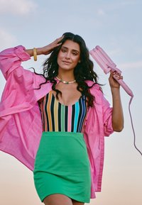 Kolorowy, pasiasty leotard w odcieniach niebieskiego, różowego i zielonego, zestawiony z zieloną spódnicą. W ręku trzymana jest różowa, oversize'owa kurtka oraz różowa lokówka.
