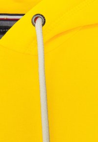 Primer plano de la tela de un sudadera amarilla con un cordón blanco pasado por un ojal plateado. Tejido de punto texturizado y costuras detalladas visibles.
