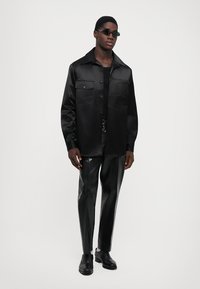 Svart satin overshirt med två bröstfickor, i kombination med glänsande svarta byxor och svarta skor, vilket skapar en elegant, monokromatisk look.