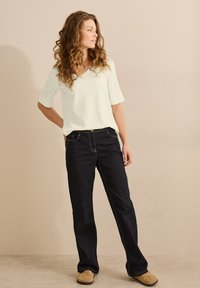 Haut vert clair à manches courtes avec un col en V, associé à un jean bleu foncé avec des coutures jaunes et une coupe décontractée. Des chaussures décontractées complètent le look.