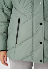 Lichtgroene donsjas met diagonale quiltstructuur, zwarte ritszak en drukknoopsluiting. Zachte, gestructureerde stof, middellange pasvorm.