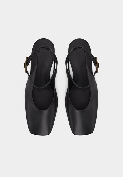 Μαύρα δερμάτινα μπαλαρίνες slingback με τετράγωνη μύτη, λεπτά λουράκια που διασταυρώνονται πάνω από το πόδι και κλεισίματα με αγκράφες σε χρυσό τόνο σε γκρι φόντο.