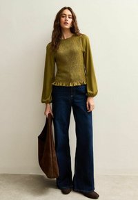 Top verde oliva arricciato con orlo a volant, abbinato a jeans svasati blu scuro. La modella tiene una borsa in suede marrone.