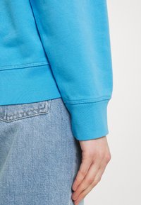 La main d'une personne reposant à côté de la poche d'un jean en denim bleu clair, portant un haut bleu vif à manches longues avec manches retroussées.