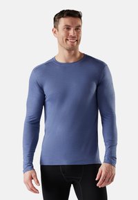 Glunderende man in een strak blauw shirt met lange mouwen en zwarte broek staat tegen een effen witte achtergrond.