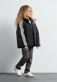 Casaco puffer preto com detalhe de três riscas brancas, combinado com calças cinzas e ténis brancos. Apresenta capuz e fecho de correr.