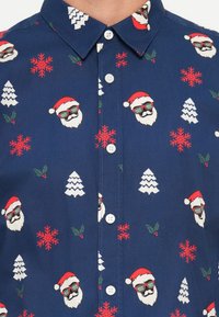 Camisa azul de botones con un patrón festivo de caras de Santa, copos de nieve, árboles de Navidad y acebo en rojo y blanco. Textura de tela suave.