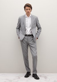 Lichtgrijs pak met een eenknops colbert, nauwsluitende pasvorm, broek met platte voorkant en een wit overhemd met knopen. Zwarte riem en schoenen.