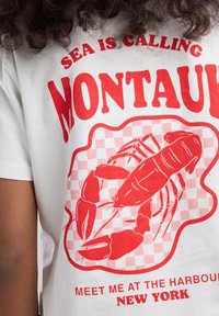 Weißes Baumwoll-T-Shirt mit rotem Grafikdruck eines Hummers auf einem karierten Hintergrund, mit dem Text "DAS MEER RUFT MONTAUK" und "TRIFF MICH IM HAFEN NEW YORK."